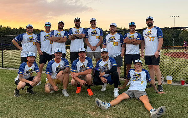 Group of softball players posing in team jerseys on a field at sunset.